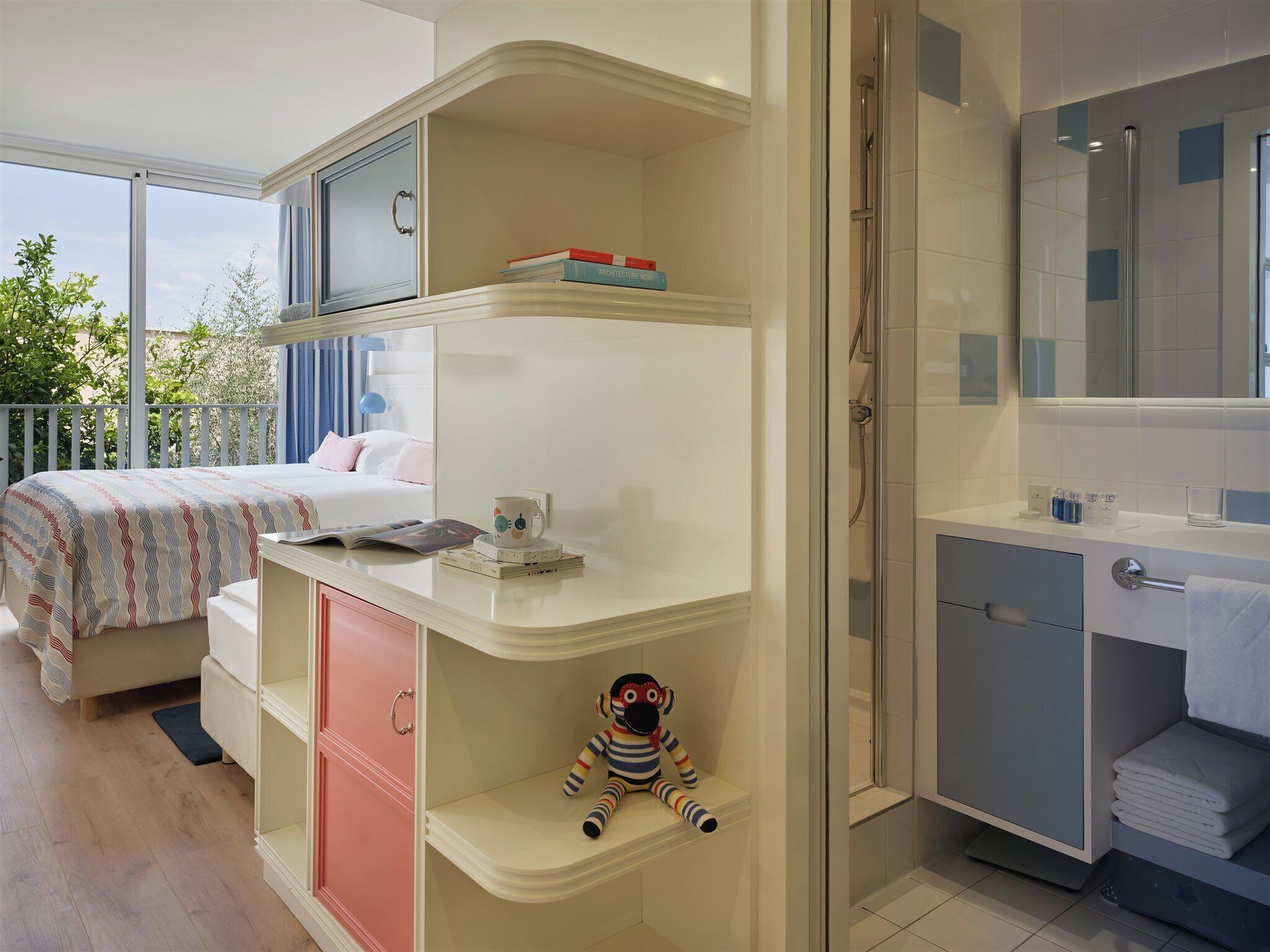 A hotel room with a bed, a desk with a chair, a small shelf with books, a plush toy, a shower area, and a bathroom with a mirror.