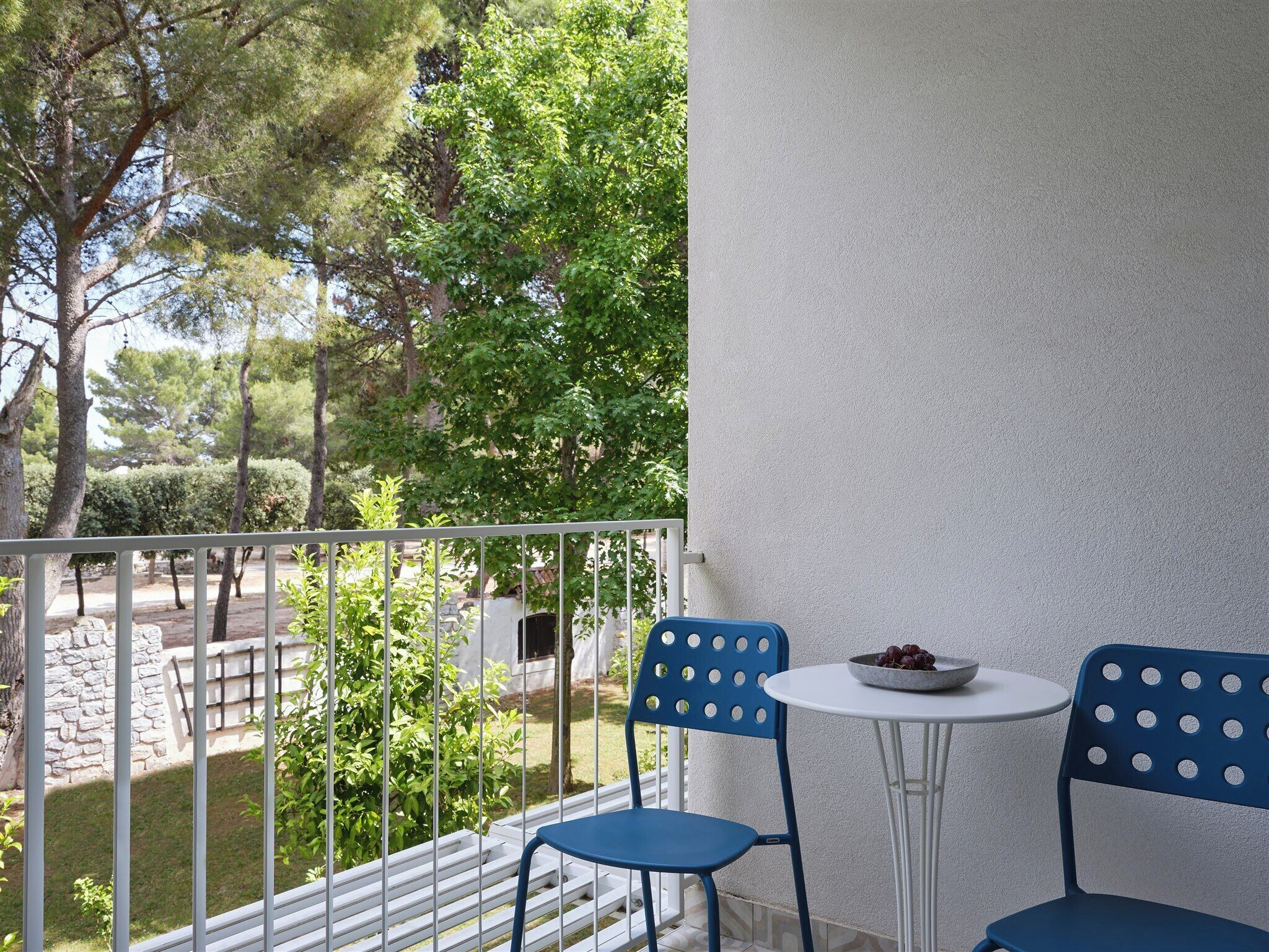 A balcony with two blue chairs and a small table, overlooking a garden with trees and a stone wall.
