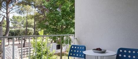A balcony with two blue chairs and a small table, overlooking a garden with trees and a stone wall.