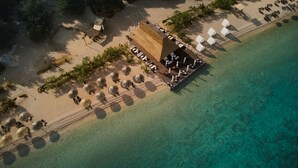 Vista aérea de un resort frente a la playa, con una estructura de techo de paja, tumbonas, sombrillas y un mar turquesa cristalino.