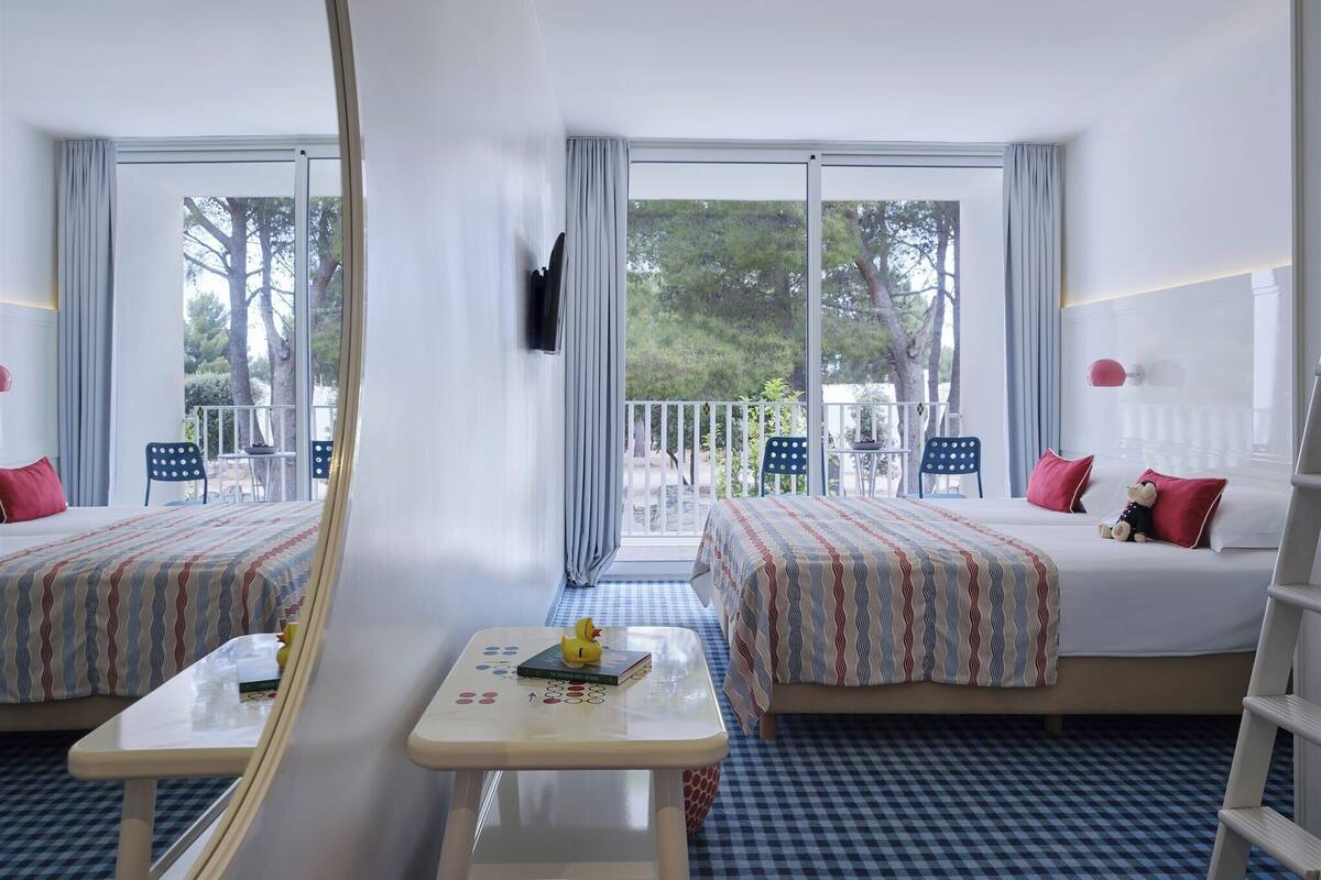 A hotel room with two beds, a small table, a chair, a TV, and a balcony with a view of trees.
