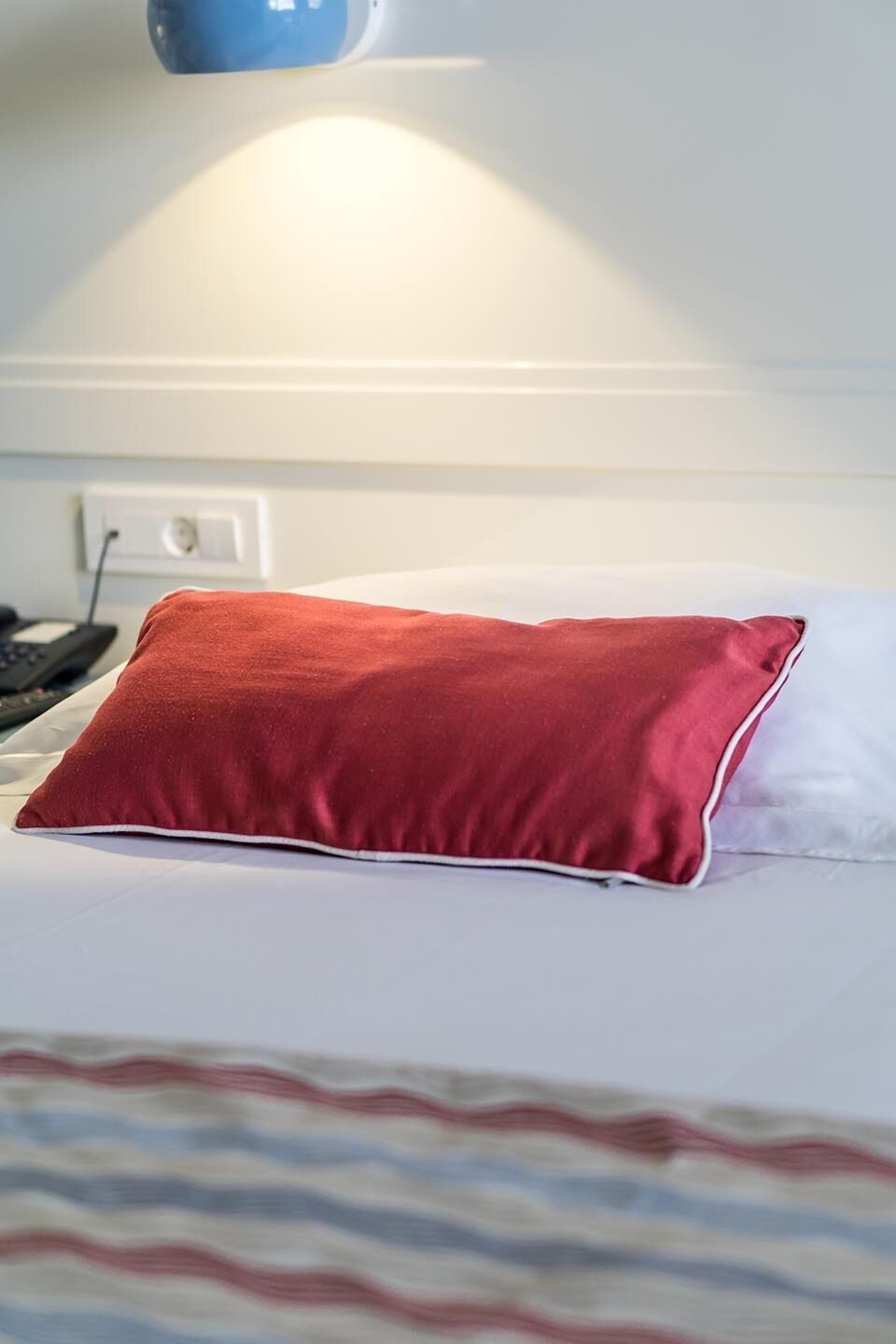 A bed with a white and red pillow, a blue lamp, and a white wall with a power outlet.
