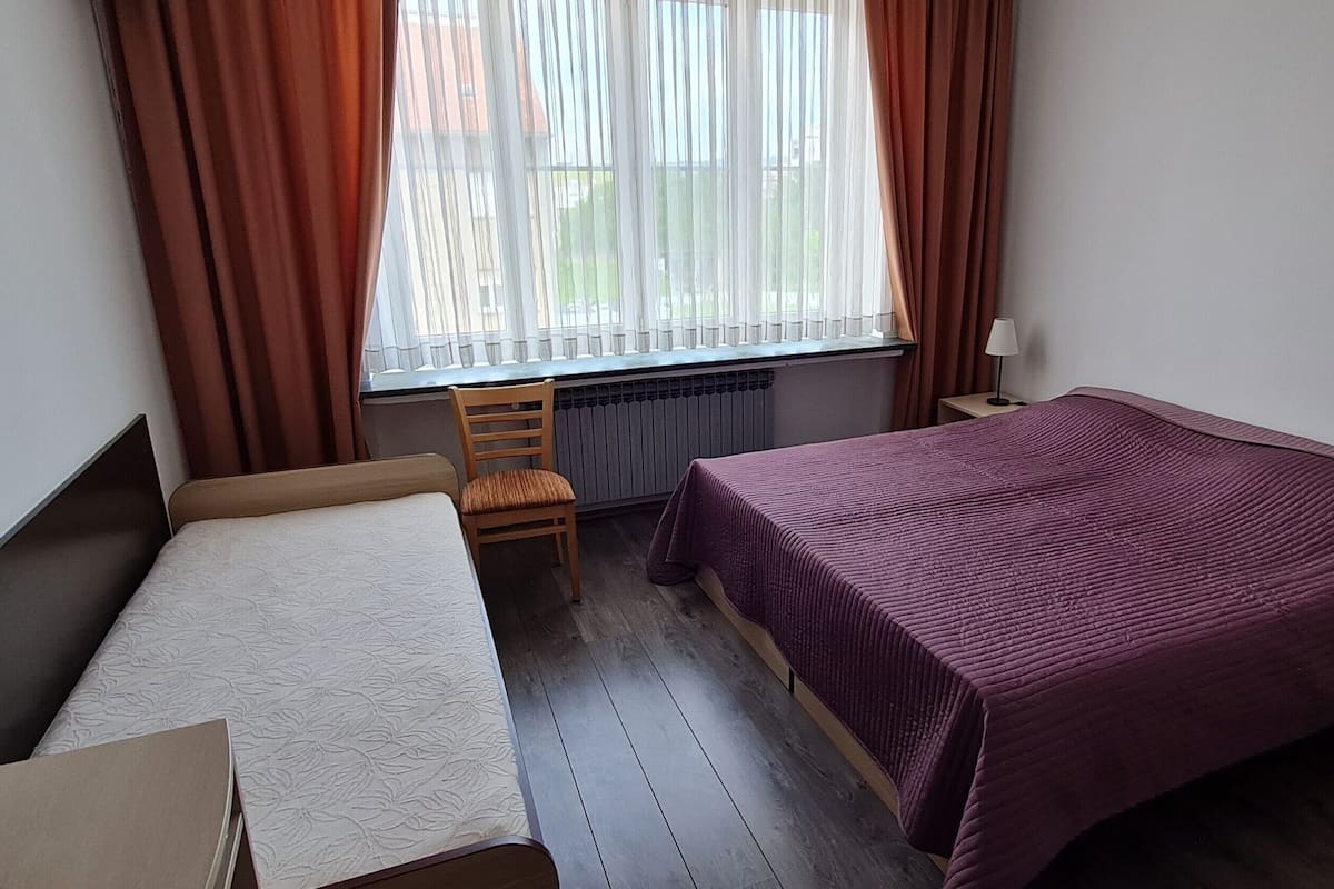 Ein Zimmer mit einem Bett, einem Sessel, einem Schreibtisch, einer Lampe und einem fenster mit Vorhängen.