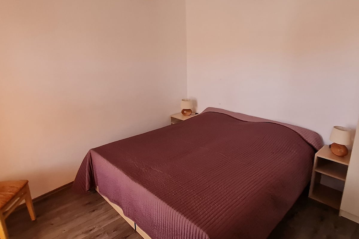 Ein Einzelbett mit lila Bettdecke, ein Nachttisch aus Holz mit einer Lampe und ein kleiner Hocker.