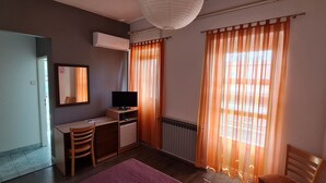 Una habitación pequeña y funcional con escritorio, silla y televisor. Incluye un radiador, un equipo de aire acondicionado y una ventana con cortinas.