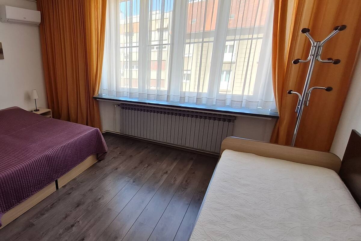 Ein Schlafzimmer mit einem Bett, einem Nachttisch, einer Lampe, einem Heizkörper und einem Fenster mit Vorhängen.