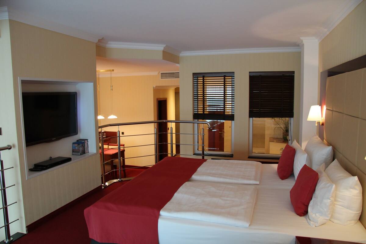 A hotel room with a bed, a TV, a desk, and a window with blinds.