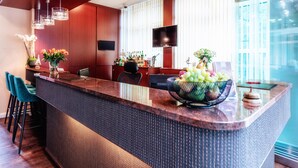 A modern bar area with a large island, bar stools, a flat-screen TV, and a large window with blinds. -  Hotel Amaris Bremerhaven (Bremerhaven)