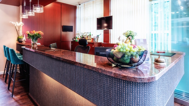 A modern bar area with a large island, bar stools, a flat-screen TV, and a large window with blinds. -  Hotel Amaris Bremerhaven (Bremerhaven)