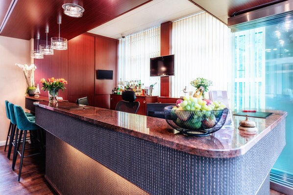 A modern bar area with a large island, bar stools, a flat-screen TV, and a large window with blinds. - Hotel Amaris (Bremerhaven)