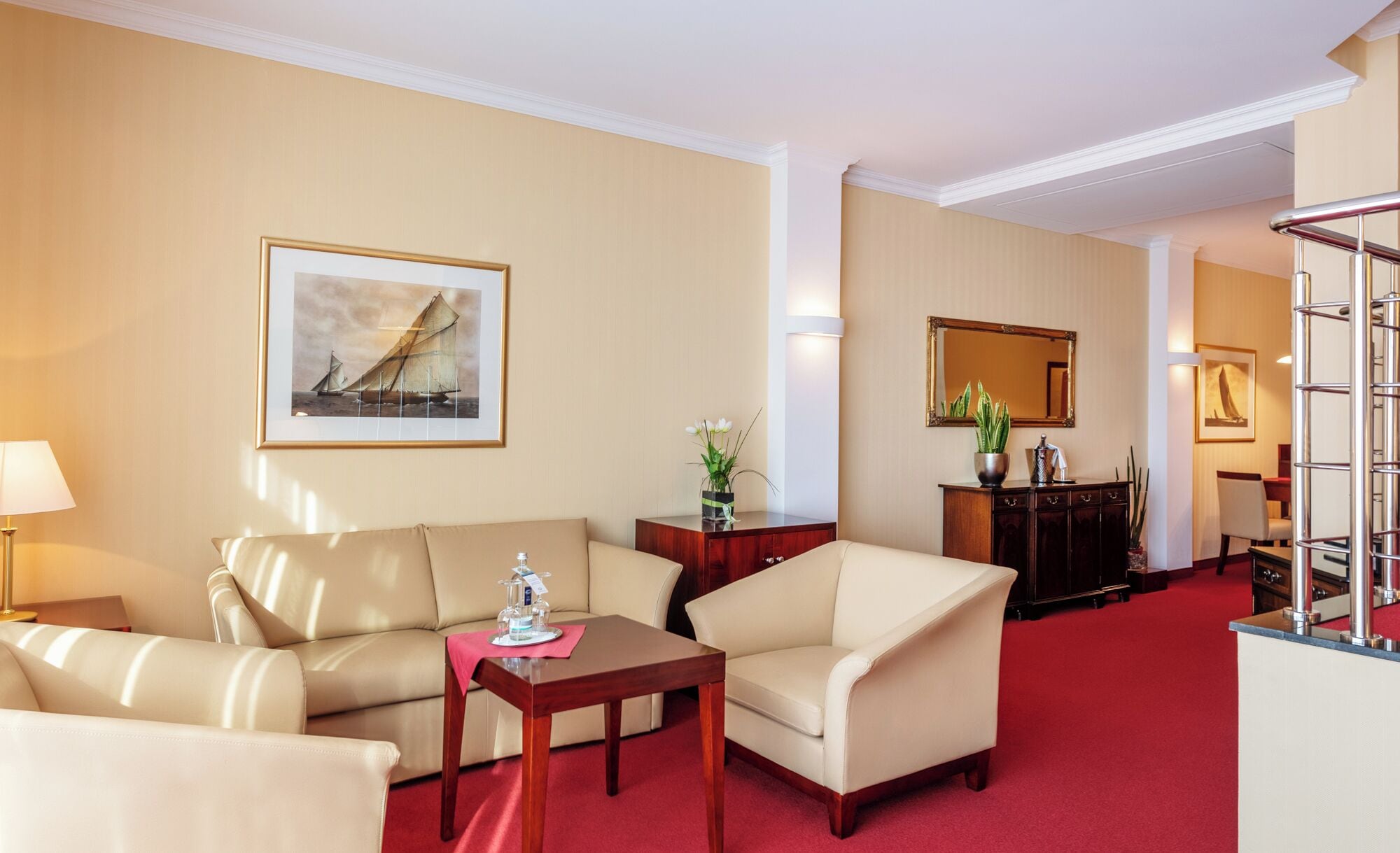 Une chambre d’hôtel avec un canapé, un fauteuil et une petite table avec une lampe. Il y a un tableau au mur et un miroir au-dessus de la commode.