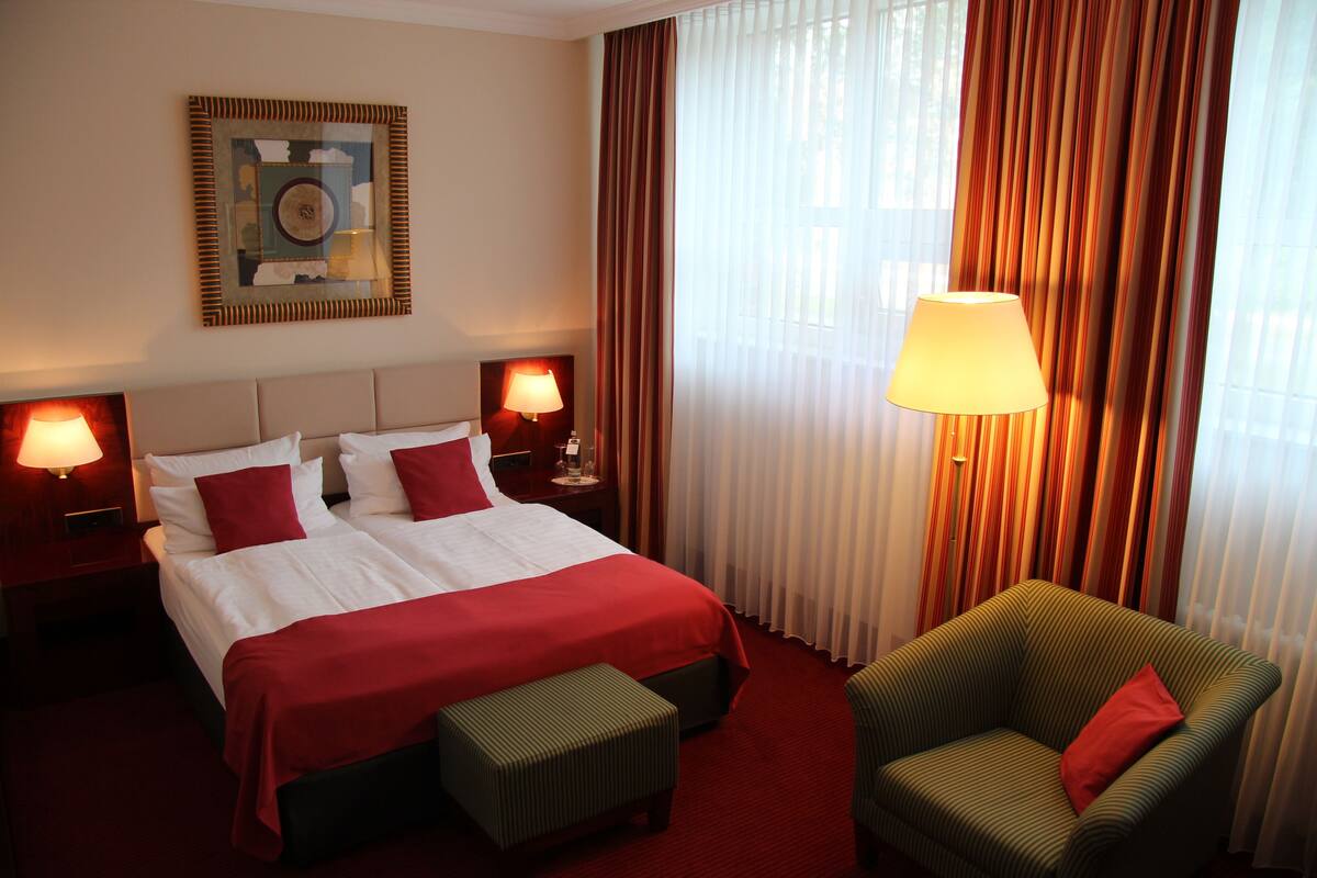 A hotel room with a large bed, two bedside lamps, a green armchair, and a window with curtains.