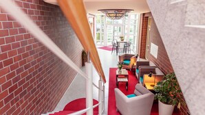 A staircase with red carpet leading to a seating area with a table and chairs, a potted plant, and a view of the outdoors. -  Hotel Amaris Bremerhaven (Bremerhaven)