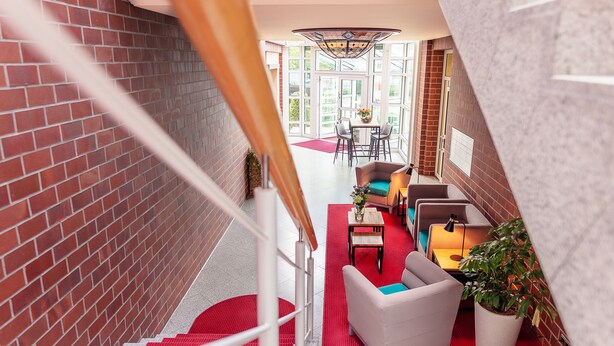 A staircase with red carpet leading to a seating area with a table and chairs, a potted plant, and a view of the outdoors. -  Hotel Amaris Bremerhaven (Bremerhaven)