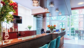 A modern hotel lobby with a reception desk, a television, and large windows offering a view of the outdoors. -  Hotel Amaris Bremerhaven (Bremerhaven)