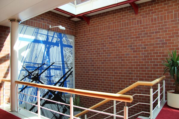 A staircase with a metal railing, a brick wall, and a large window with a blue abstract painting. - Hotel Amaris Bremerhaven (Bremerhaven)