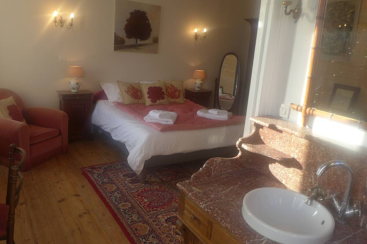 A bedroom with a bed, a chair, a nightstand, a mirror, and a bathroom with a sink and mirror.