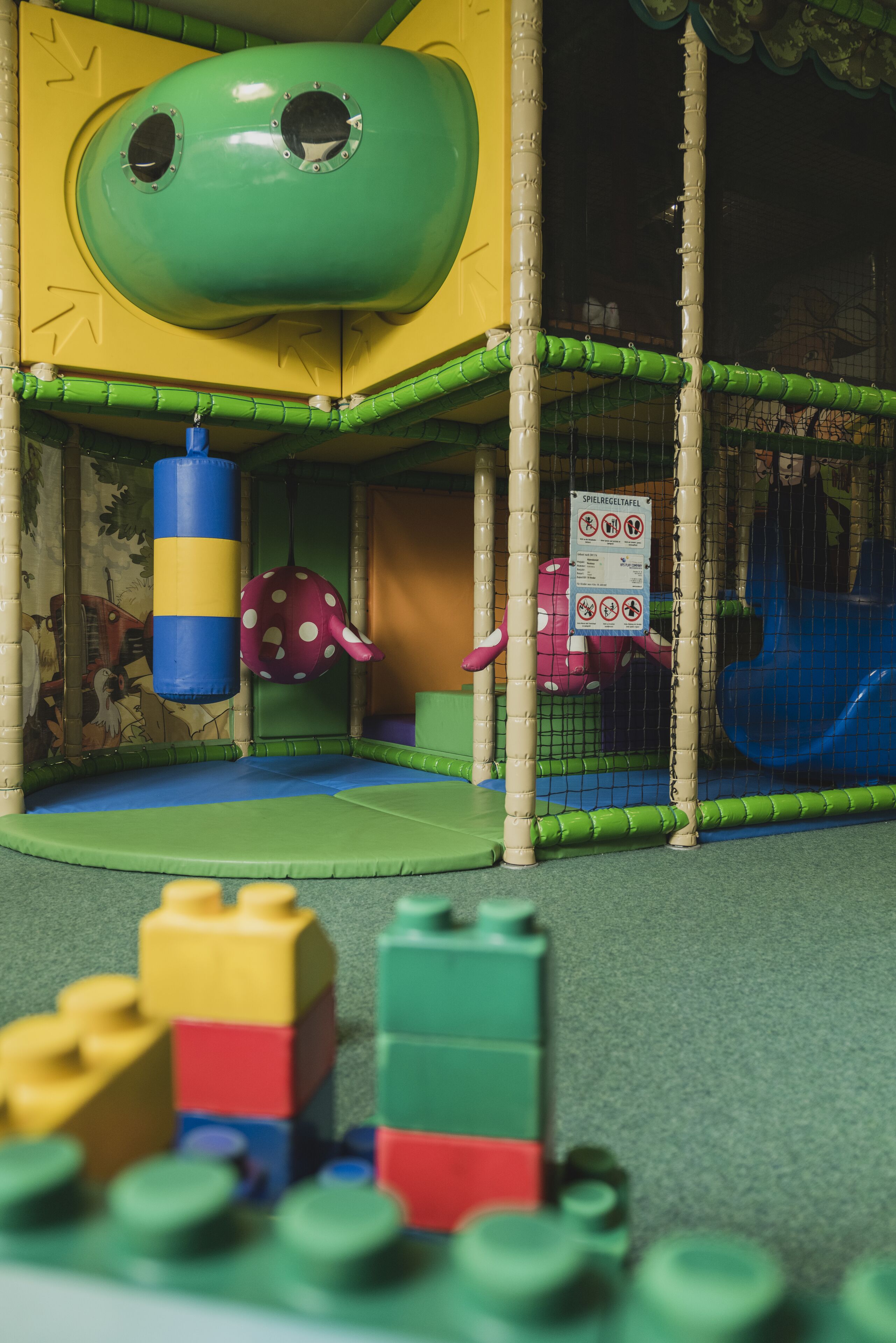 A children's play area with a green and yellow play structure, a blue slide, and a soft play floor.