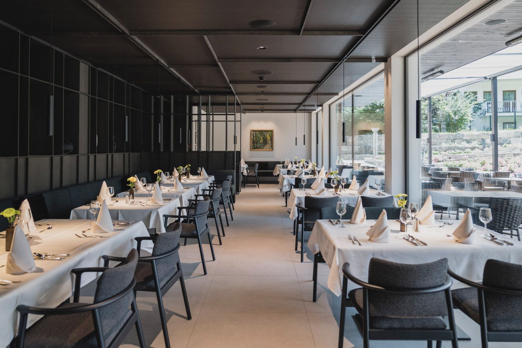 A restaurant with tables set for dining, a large window on the right, and a painting on the wall.