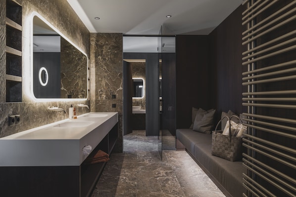 A modern bathroom with a large mirror, two sinks, a walk-in shower, and a seating area with a towel and a handbag. - Neuhaus Zillertal Resort (Mayrhofen)