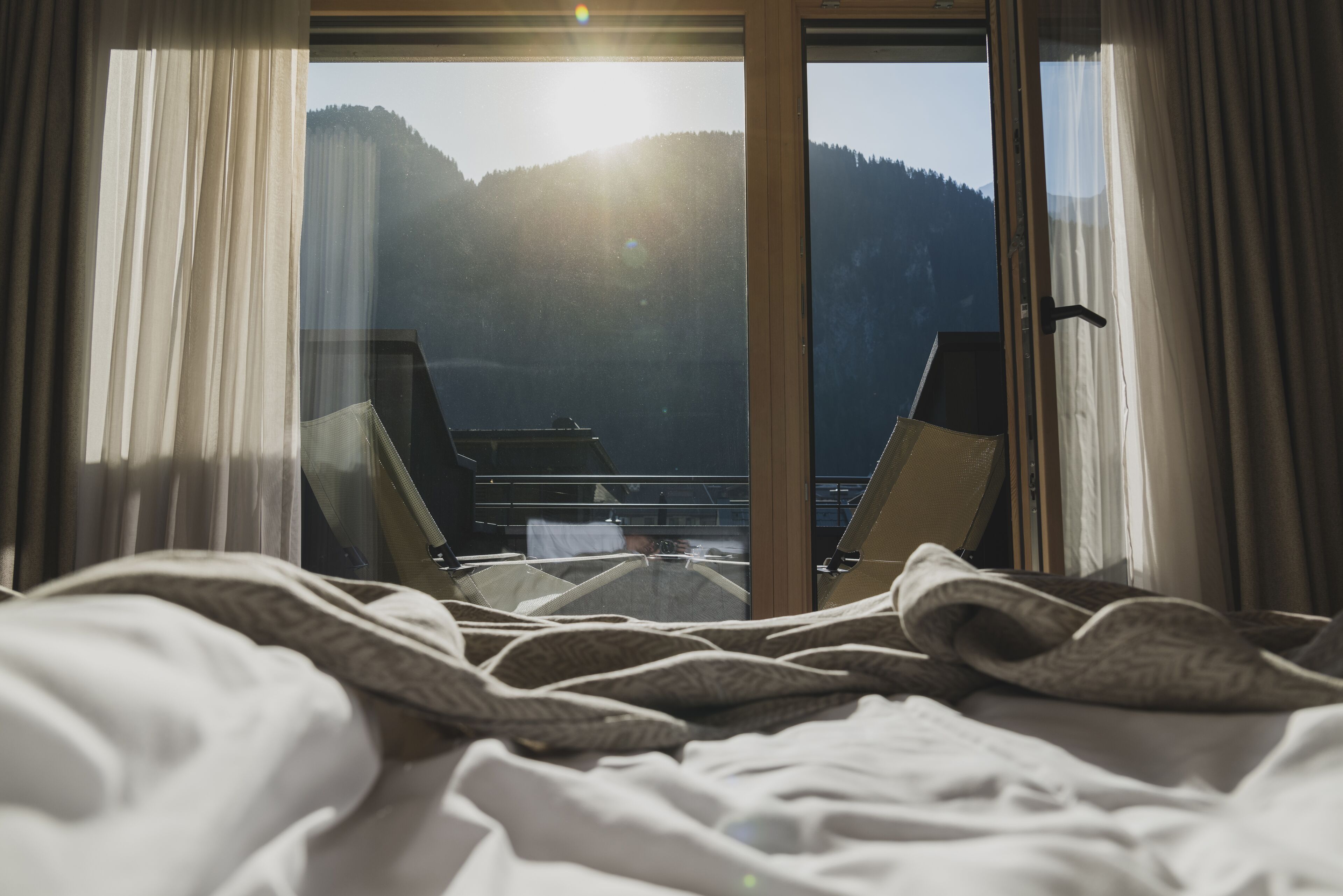 A hotel room with a bed, a view of mountains through a large window, and a balcony with chairs.