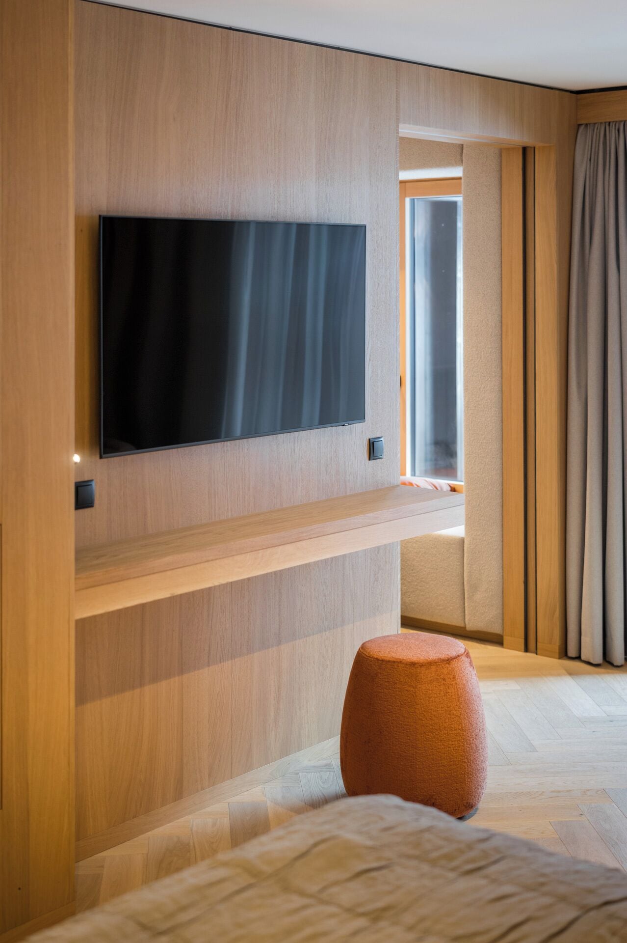 Ein modernes Hotelzimmer mit einem Flachbildfernseher, der an einem Holzpaneel befestigt ist, einem kleinen orangefarbenen Hocker und einem großen Fenster mit Vorhängen.