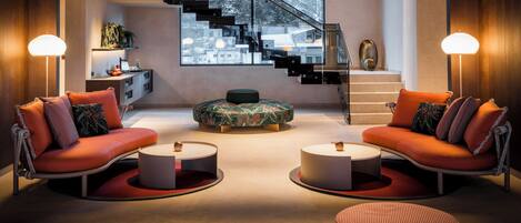 Una sala de estar moderna con dos sofás, una mesa de centro y un otoman redondo. Se ve una escalera con barandilla y vistas al exterior.