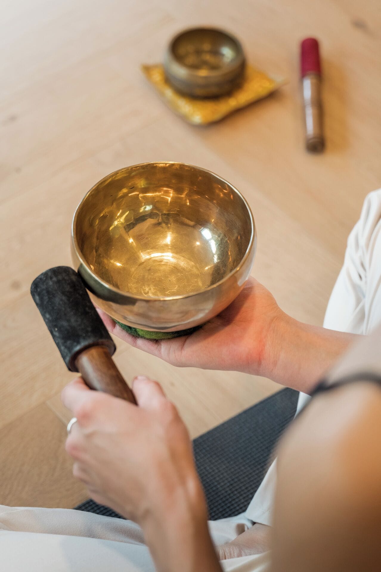 Une personne tenant un bol en laiton, un maillet noir et un objet rouge sur un plancher en bois.
