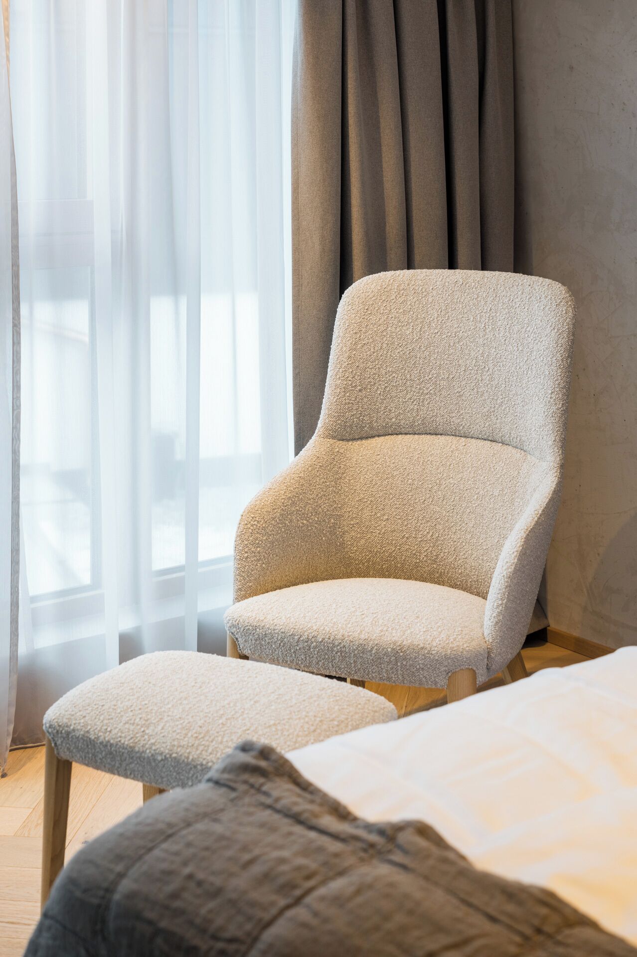 A room with a light-colored armchair and ottoman, a bed with a white bedspread, and a window with sheer curtains.