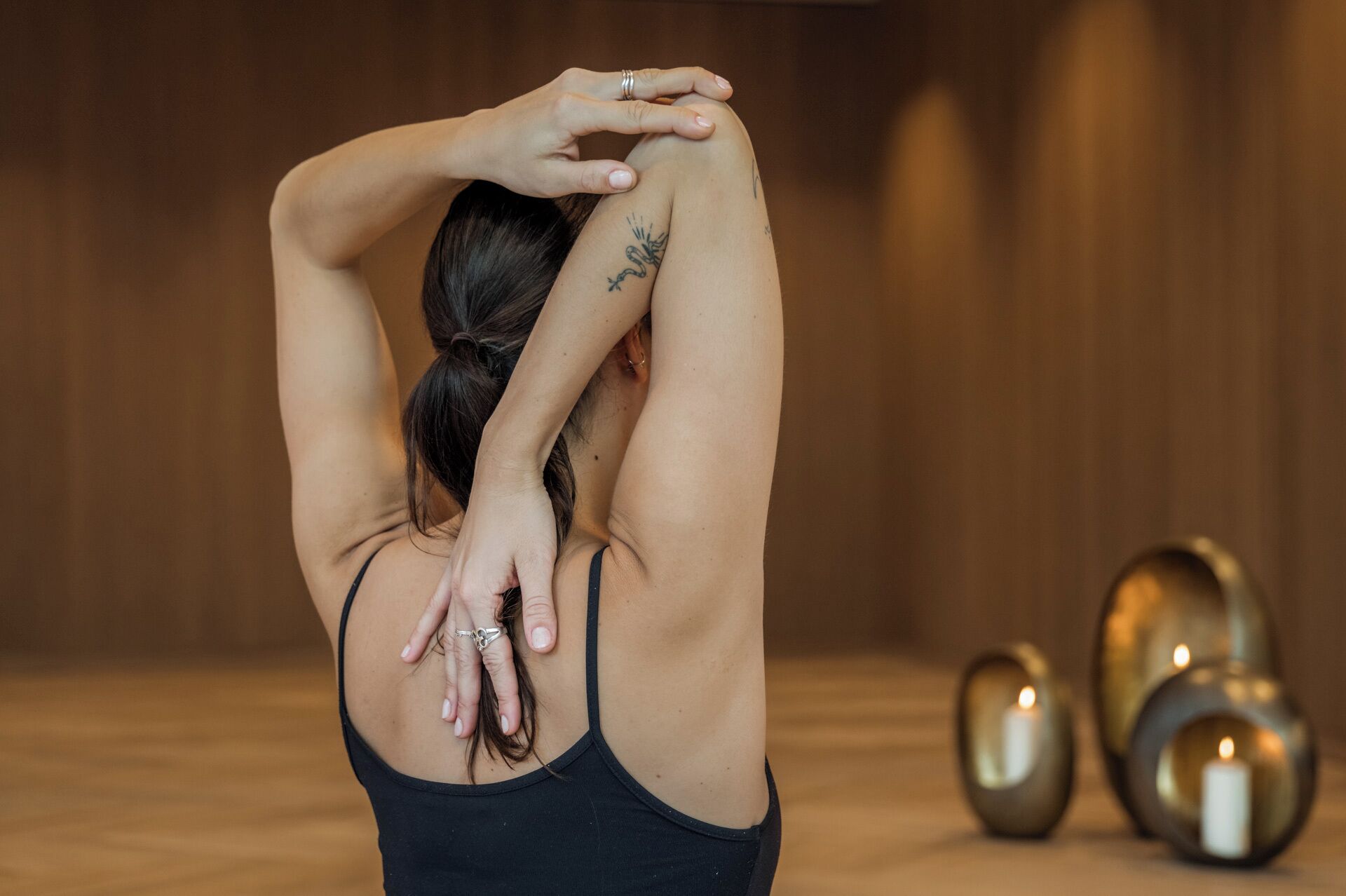 A person in a room with wooden walls and a floor, wearing a black tank top. There are candles and metallic objects on the floor.
