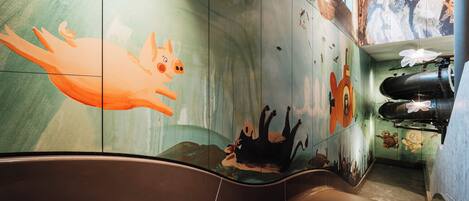 Un baño moderno con una bañera grande, un mural con una rana y un perro, y una zona de ducha con una manguera negra.