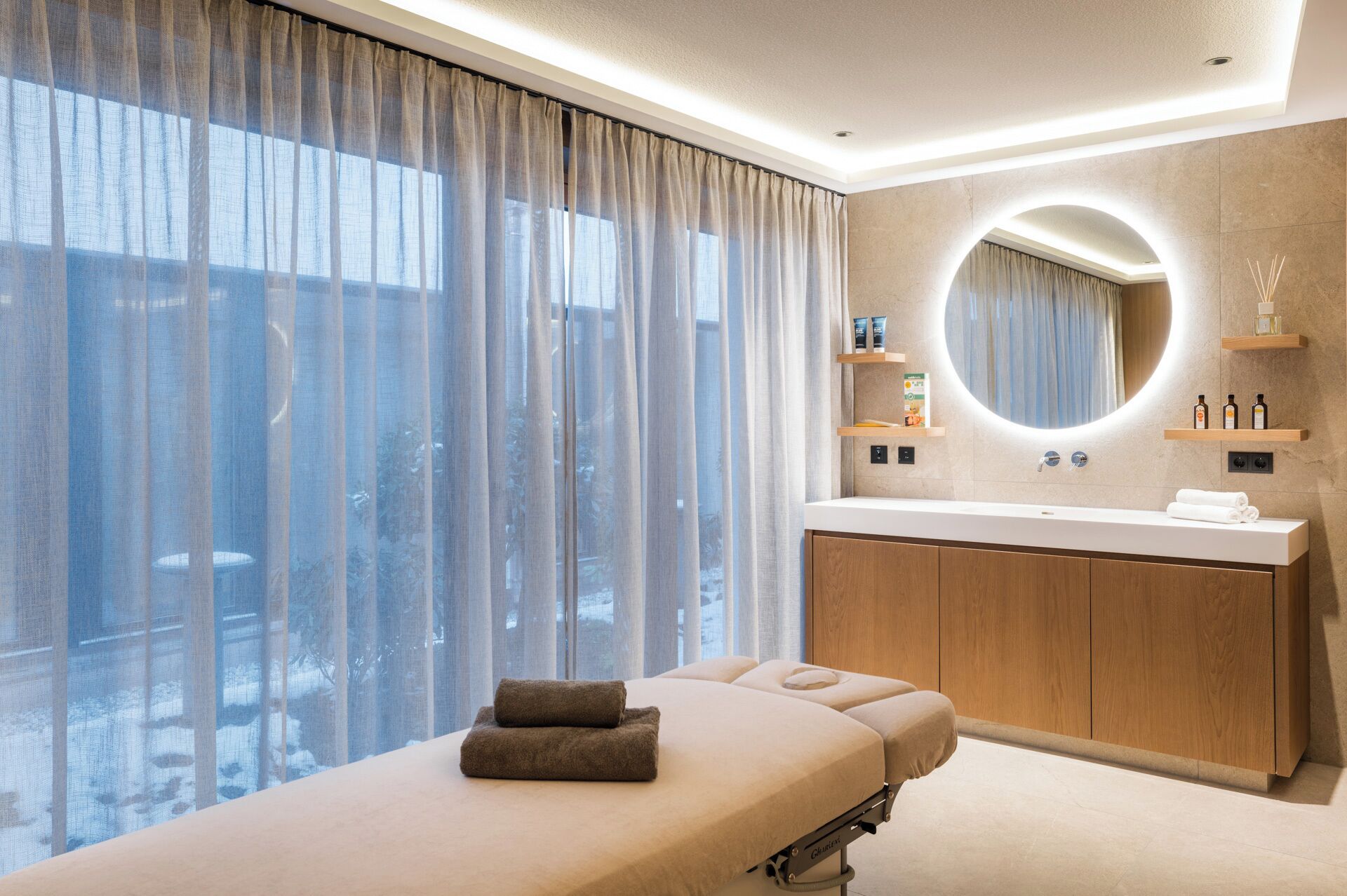 Una habitación de hotel moderna con un gran ventanal, una camilla de masaje, un espejo redondo y un tocador de madera.