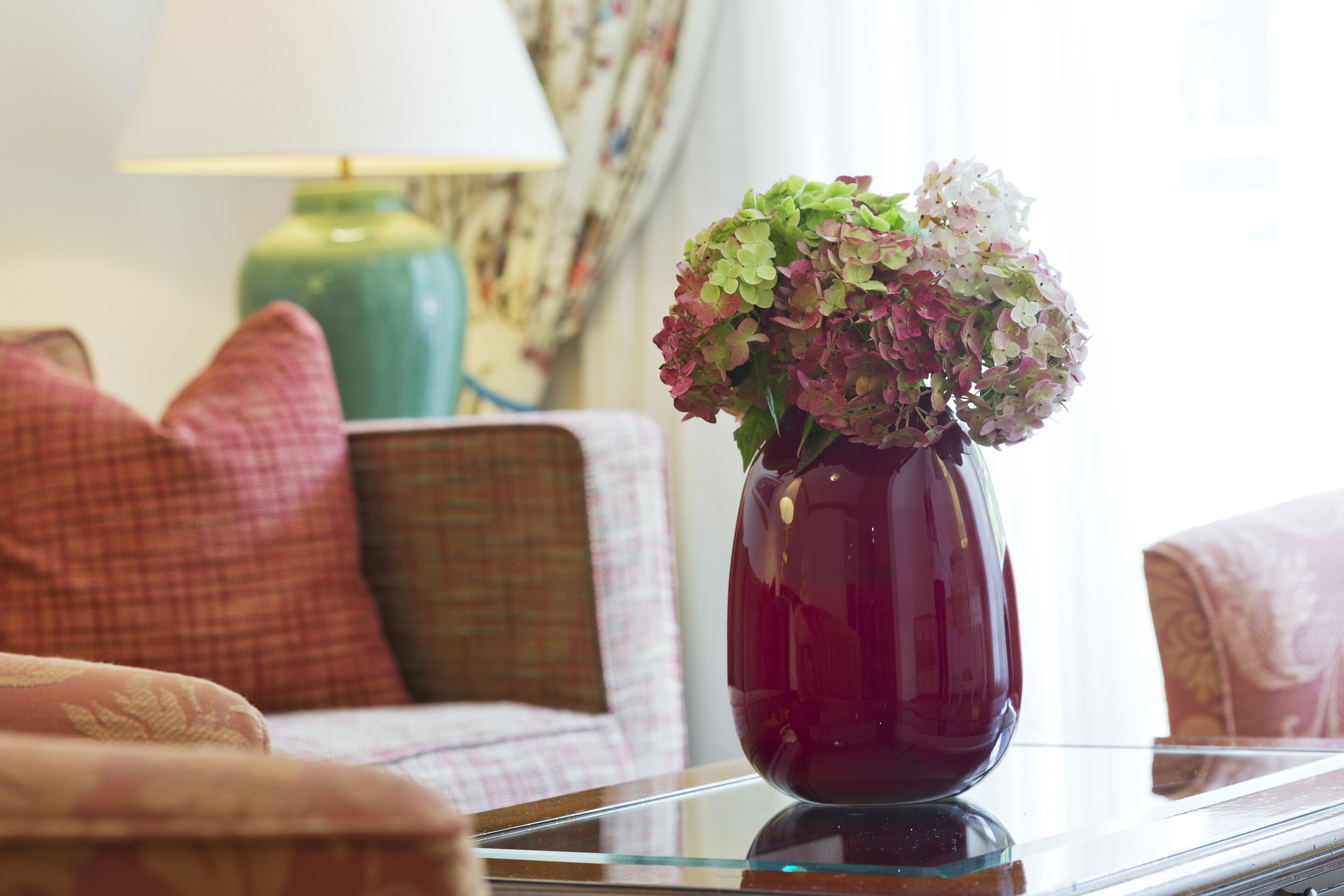 A vase with flowers on a glass table in a room with a lamp and patterned curtains.