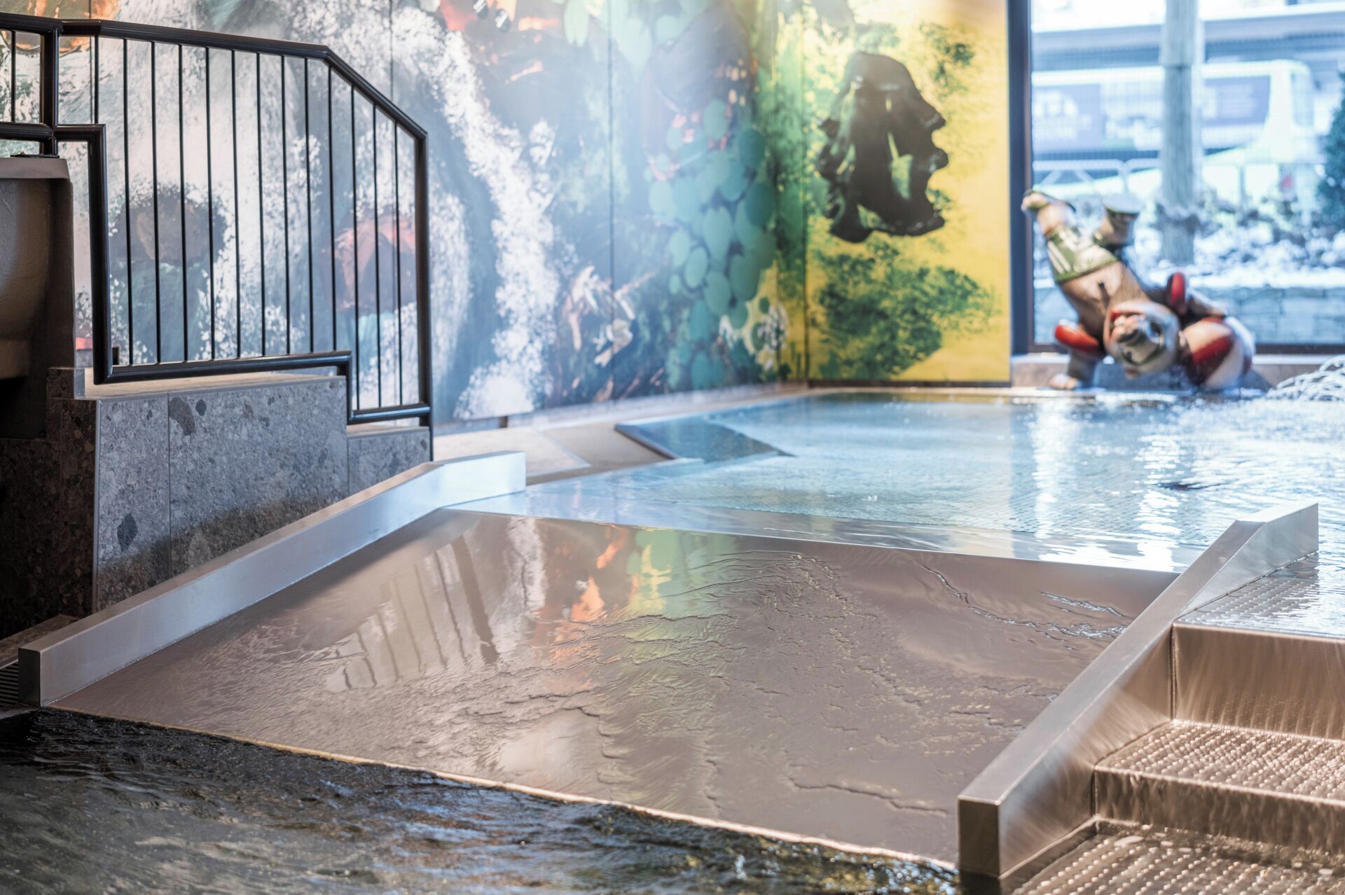 An indoor swimming pool with a modern design, featuring a clear water surface, a metal staircase, and a mural on the wall.