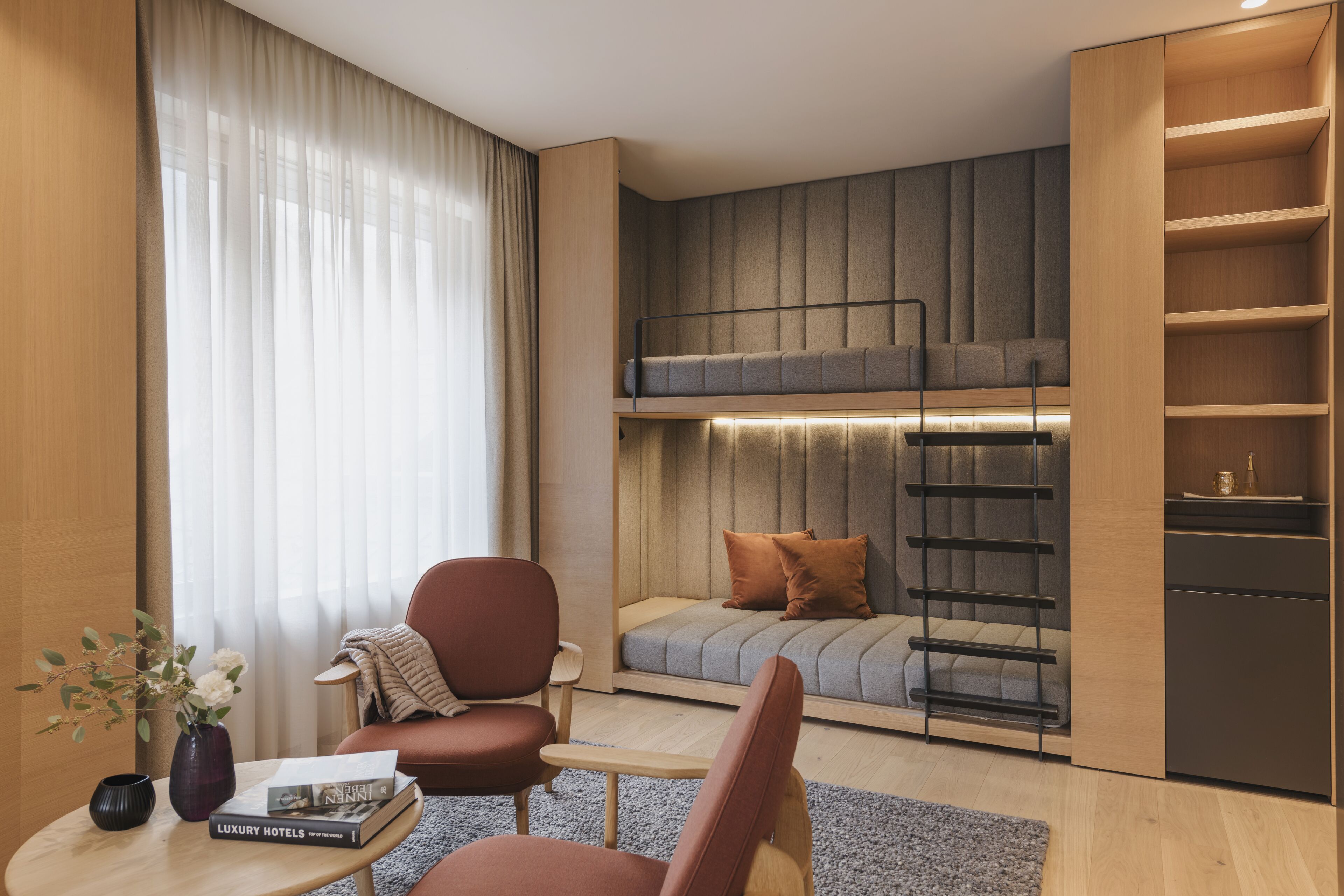 A modern hotel room with a bunk bed, a sofa, a coffee table with books, and a vase with flowers.