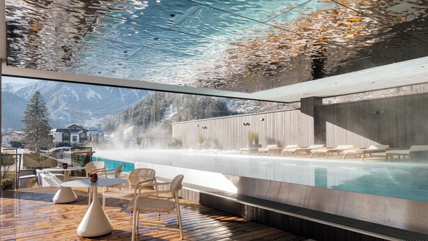 Un espace piscine extérieur moderne offrant une vue sur les montagnes, avec une terrasse en bois, des chaises longues et un toit de verre qui reflète le ciel.