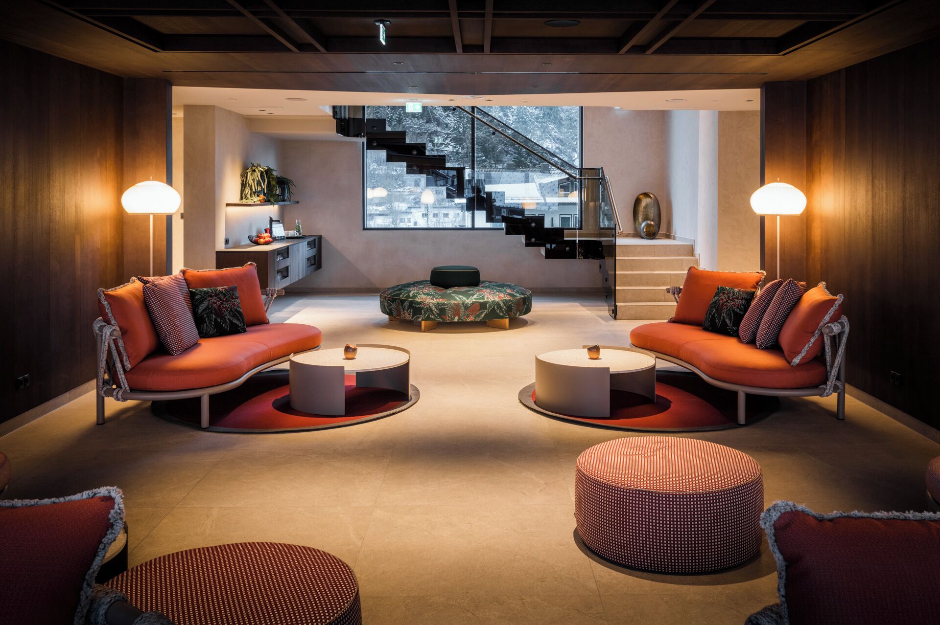 A modern living room with two sofas, a coffee table, and a round ottoman. A staircase is visible in the background.
