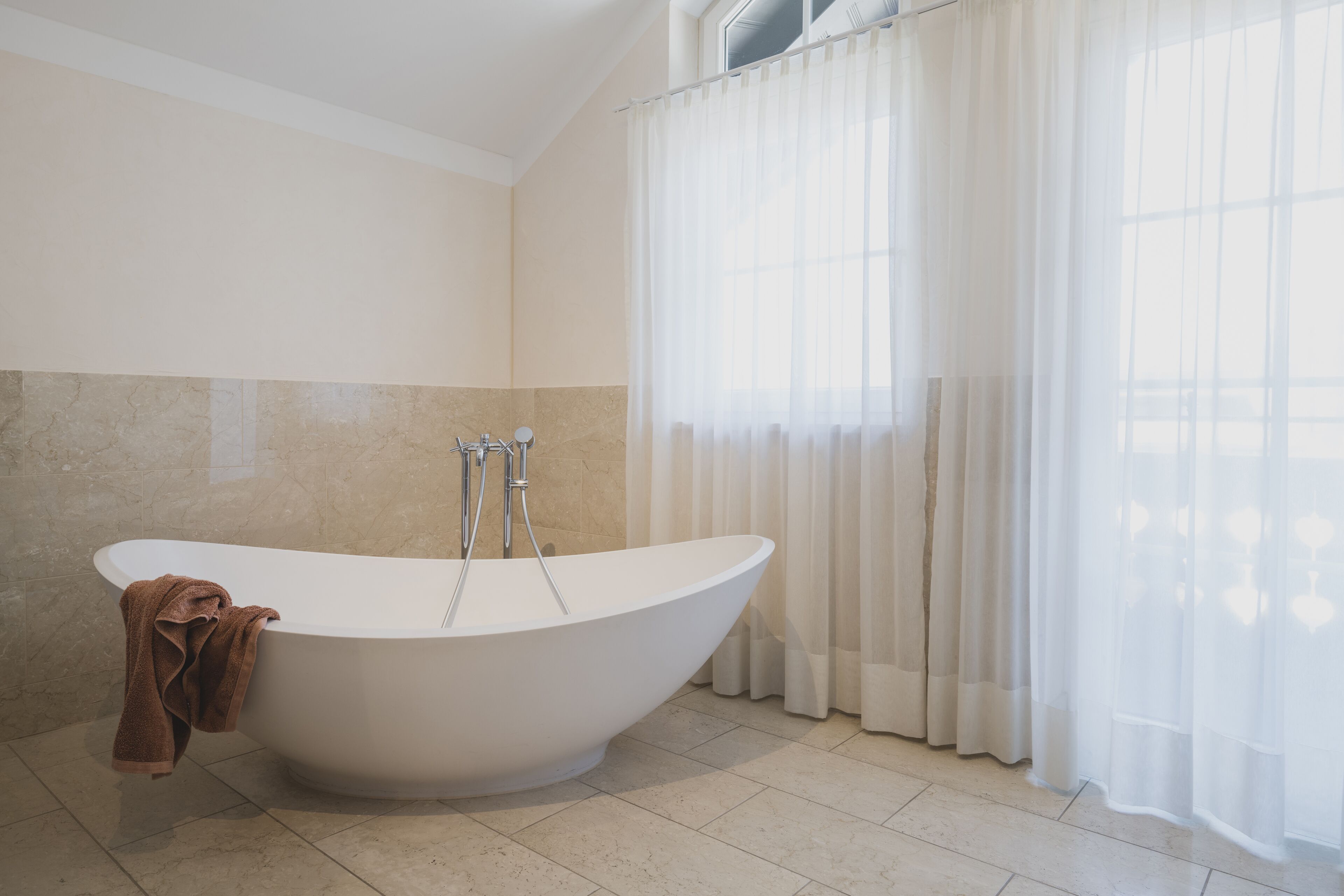 A modern bathroom with a freestanding bathtub, a towel, and a window with sheer curtains.