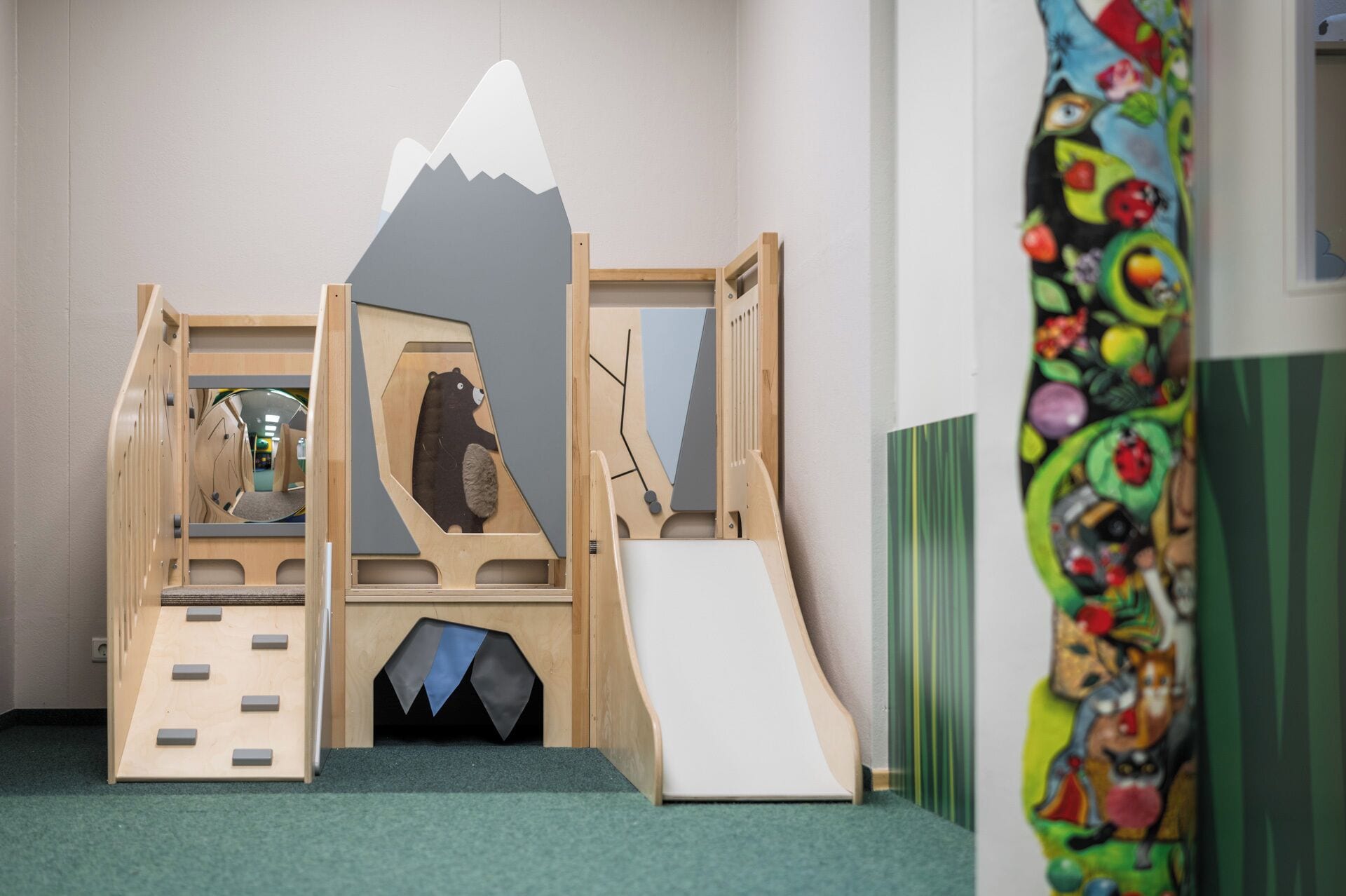 A children's play area with a wooden climbing structure, a slide, and a colorful wall mural.