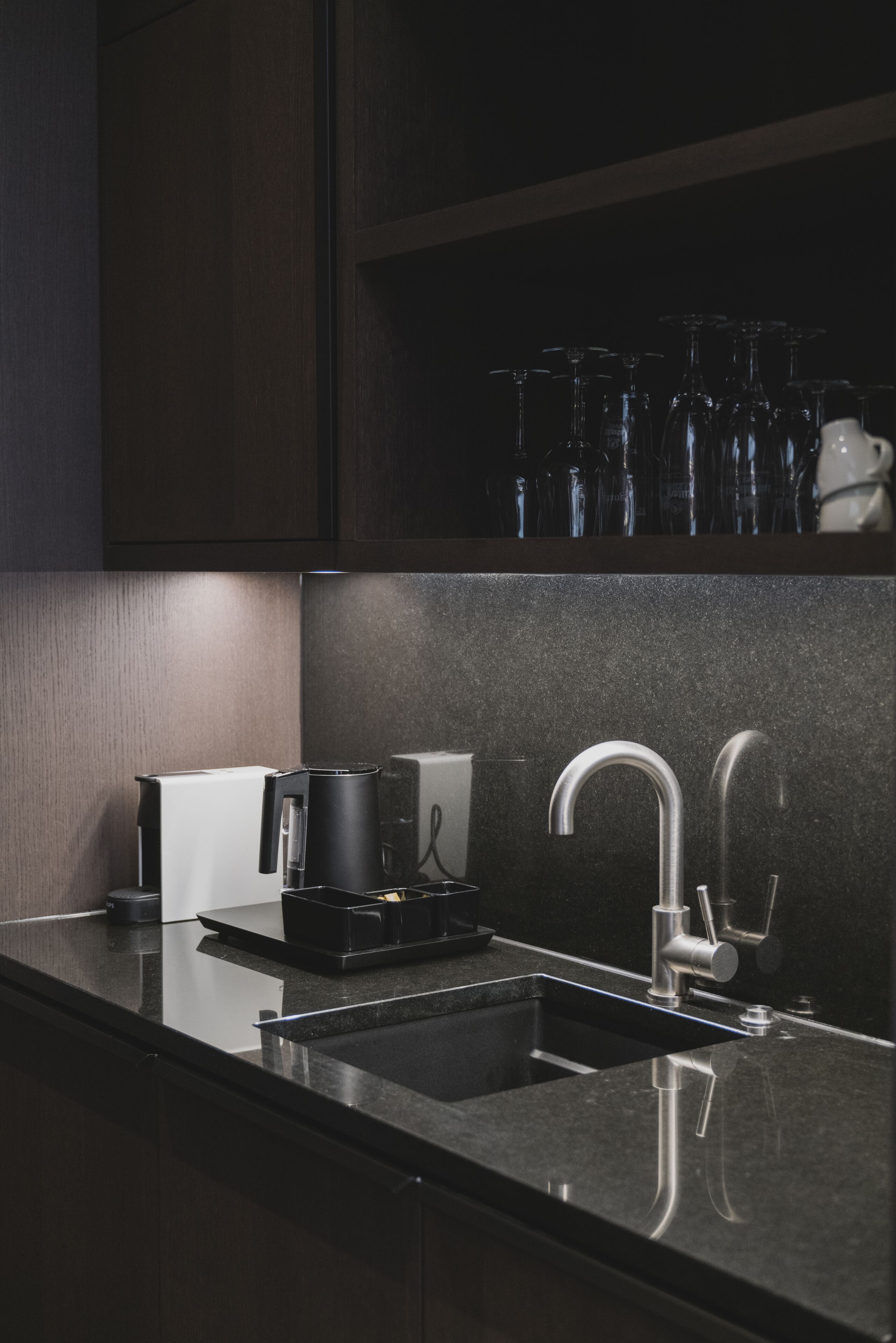 Uma cozinha moderna com bancada escura, pia de aço inoxidável e cafeteira.