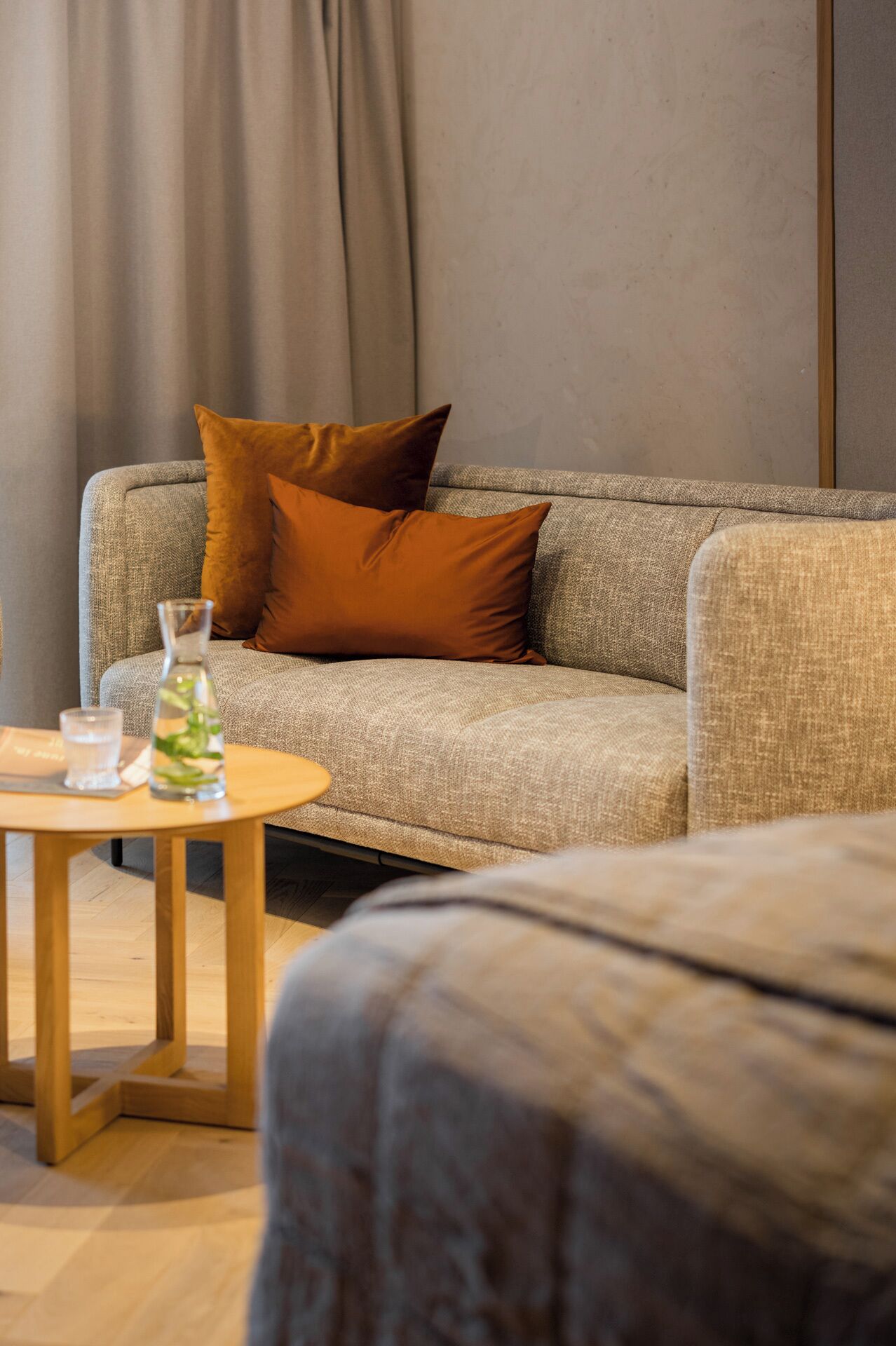 Un salon moderne avec un canapé gris, deux coussins marron, une petite table en bois sur laquelle se trouvent un verre d’eau et une bouteille transparente.