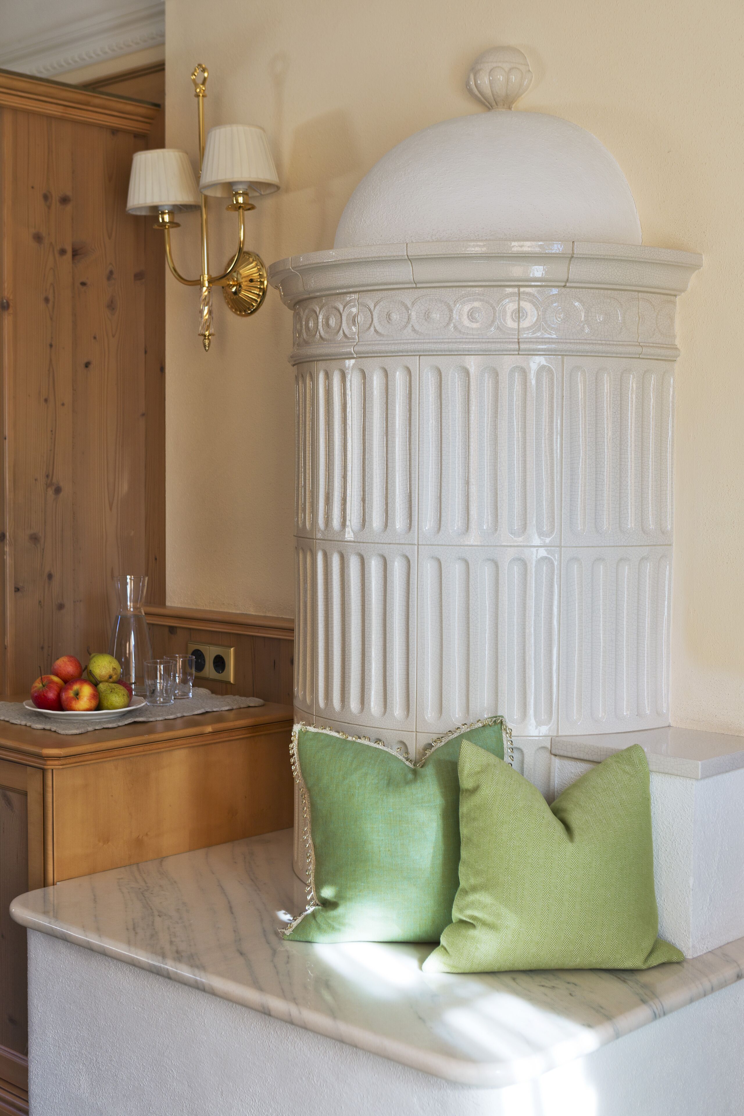 A marble countertop with a white decorative urn, two green cushions, and a wooden sideboard with fruits and glasses.