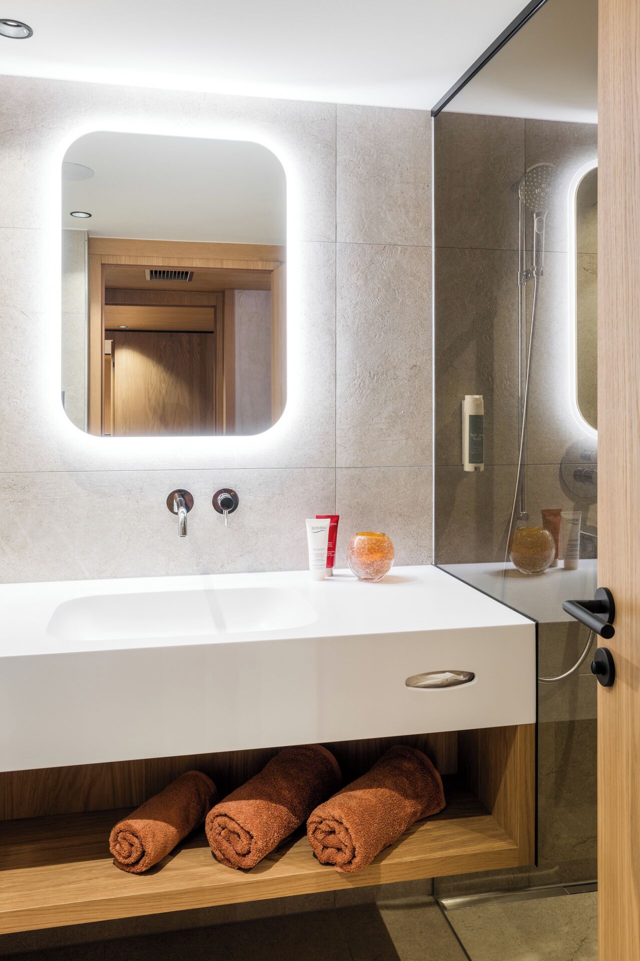 Een moderne badkamer met een witte wastafel, twee kraanwerkjes, een spiegel en een douchecabine met glazen wanden. Op het aanrecht liggen handdoeken en toiletartikelen.