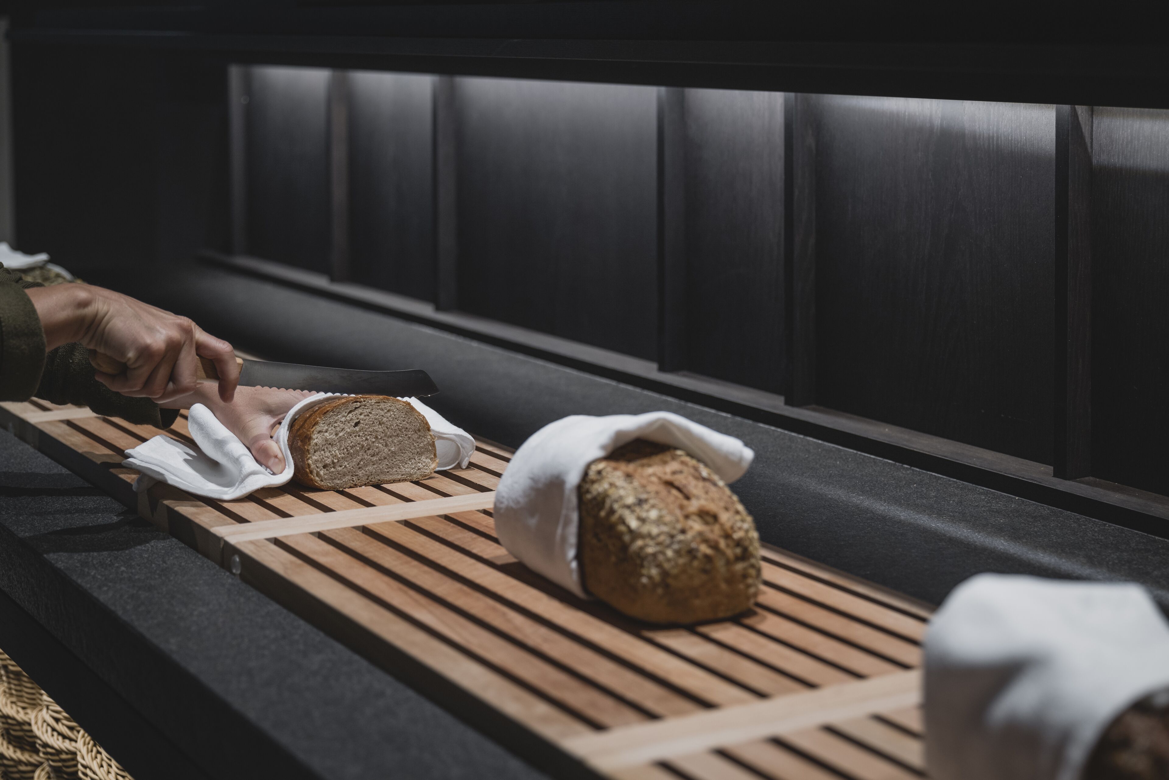 A wooden tray with bread and a loaf wrapped in a cloth, placed on a bench in a dimly lit room.