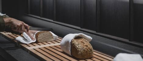Ein hölzernes Tablett mit Brot und einem in Stoff gewickelten Laib, platziert auf einer Bank in einem schummrigen Raum.