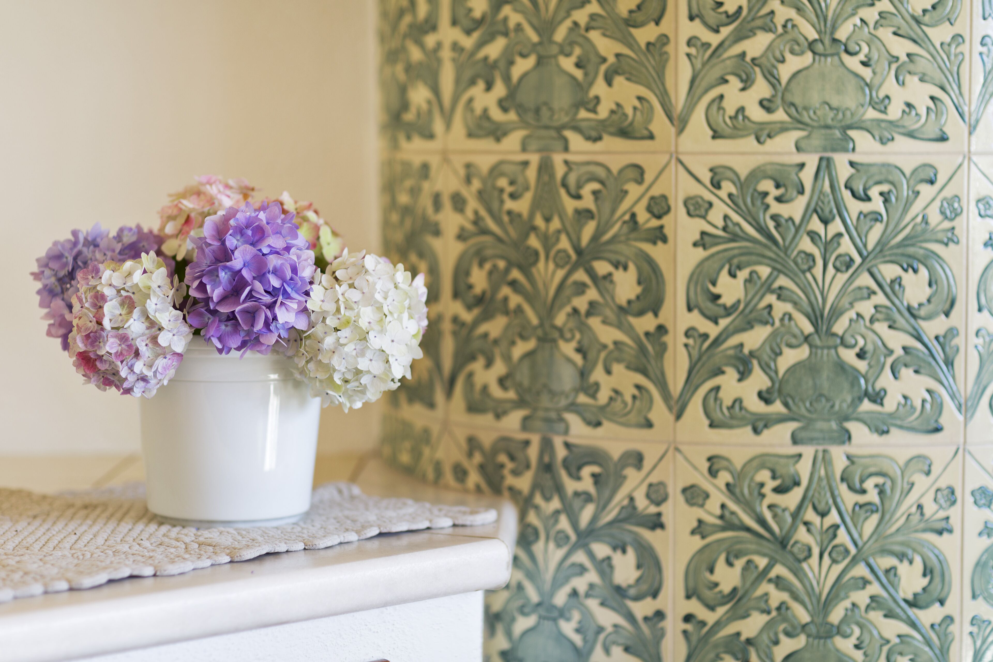 Eine weiße Vase mit Blumenarrangement auf einer weißen Oberfläche, im Hintergrund eine Wand mit gemusterten Fliesen.