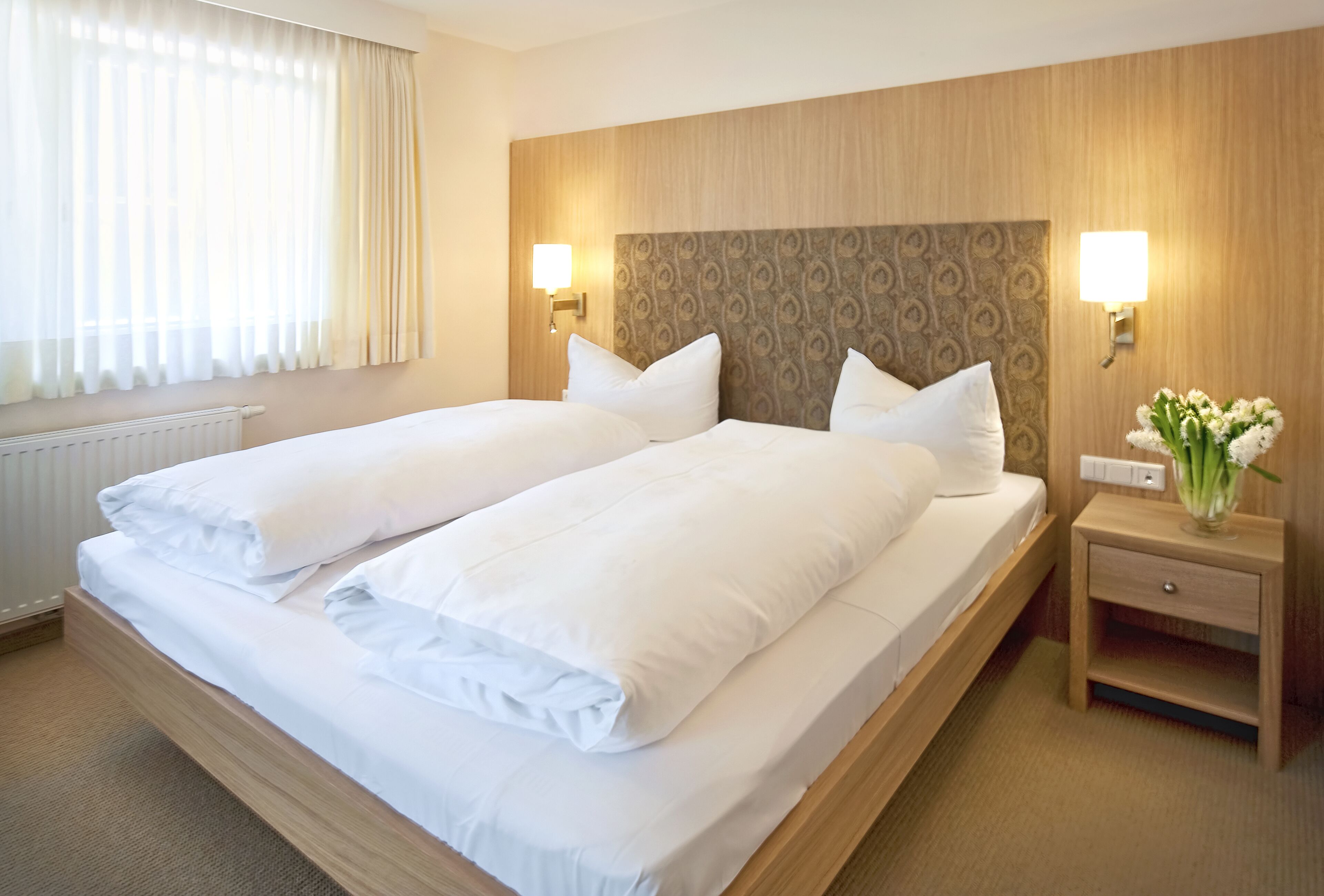 A hotel room with two single beds, a wooden headboard, bedside lamps, a nightstand with a vase of flowers, and a radiator.