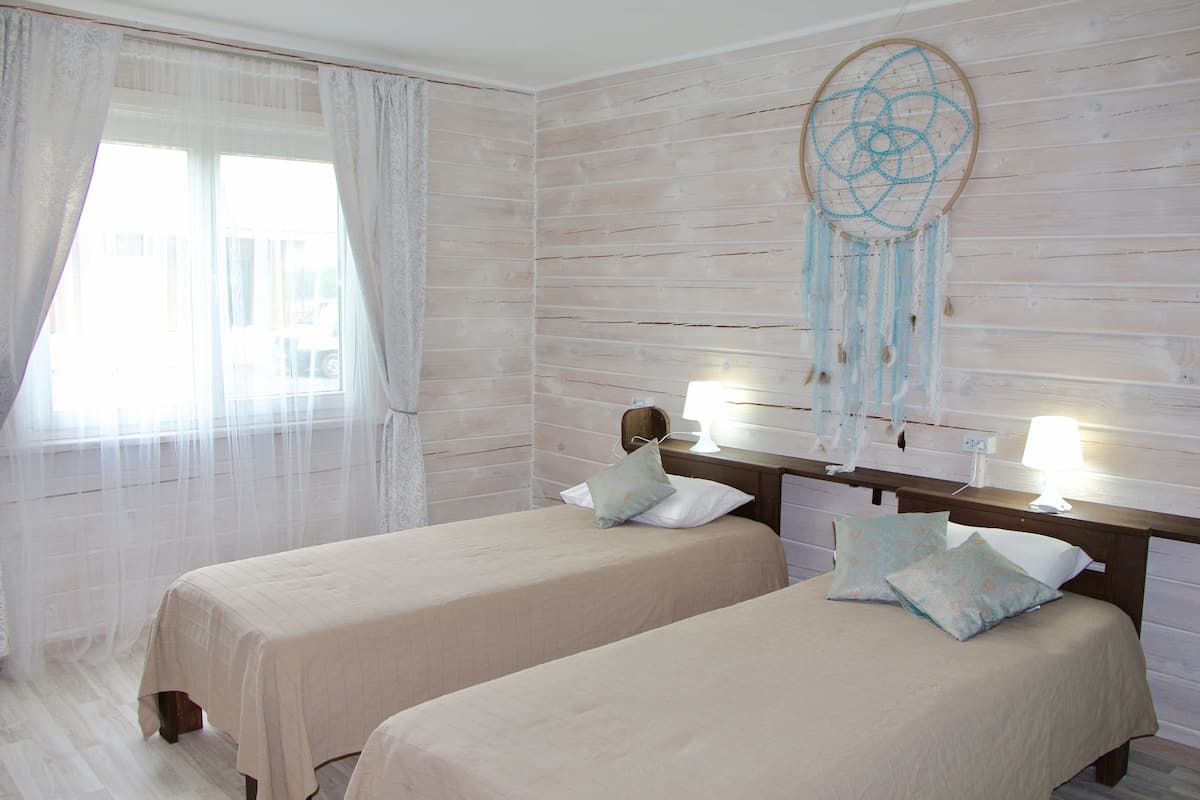 A room with two beds, a large window with sheer curtains, a dreamcatcher on the wall, and a bedside lamp.