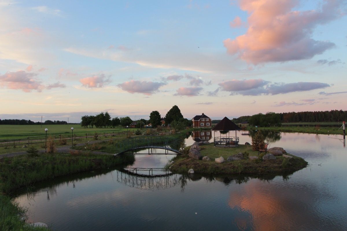 A serene lakeside setting with a wooden bridge, a gazebo, and a house with a red roof.