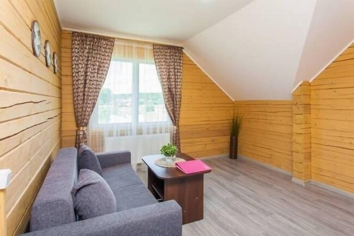 A cozy room with a wooden slatted ceiling, a grey sofa, a small wooden table with a pink folder, and a window with patterned curtains.